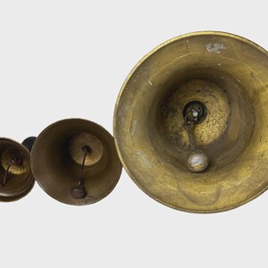 May include: Three antique brass bells of varying sizes are arranged in a row, viewed from above. The bells have a tarnished, aged appearance, with visible wear and tear. Each bell features a clapper and a loop for hanging, suggesting a decorative or functional purpose.