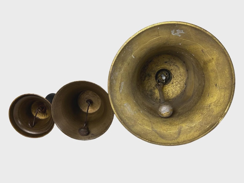 May include: Three antique brass bells of varying sizes are arranged in a row, viewed from above. The bells have a tarnished, aged appearance, with visible wear and tear. Each bell features a clapper and a loop for hanging, suggesting a decorative or functional purpose.