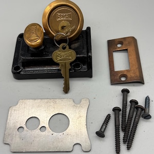May include: An antique Segal lock set with a brass-colored knob and lock face, a key, and a black metal base. Includes a metal strike plate, a metal plate, and several screws. The lock set is designed for a door.