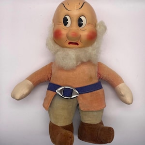 May include: Vintage plush doll of Grumpy from Snow White. The doll features a bald head, rosy cheeks, and a white beard. He wears a peach-colored tunic, blue belt with a silver buckle, and brown boots. The doll's arms and hands are a lighter peach color.