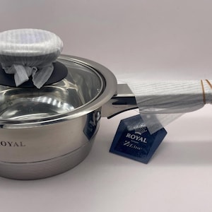May include: Stainless steel saucepan with a glass lid and long handle. The pan is wrapped in protective packaging. A tag with the text "ROYAL Z Line" is attached to the handle.
