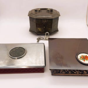 May include: Three antique boxes are displayed. A tarnished silver rectangular box with a circular emblem, a dark brown box with a floral emblem, and a bronze-colored octagonal box with a lock and key.