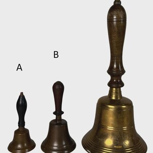 May include: Three antique brass bells with wooden handles. The bells vary in size, with the largest bell featuring a long, ornate handle. The smallest bell has a black handle. The bells are displayed against a neutral background.