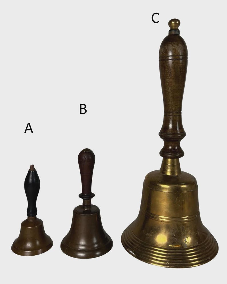 May include: Three antique brass bells with wooden handles. The bells vary in size, with the largest bell featuring a long, ornate handle. The smallest bell has a black handle. The bells are displayed against a neutral background.