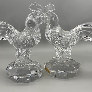 May include: Two clear crystal rooster figurines. The roosters are facing each other, with their beaks almost touching. Each rooster has a detailed, textured body, a comb, and a tail. They are mounted on faceted hexagonal bases.