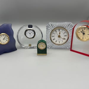 May include: A collection of five decorative quartz clocks. The clocks feature various shapes and materials, including a blue wavy clock, a clear crystal clock, a green clock, a square crystal clock, and a red clock. Each clock has a gold-tone bezel and Roman numeral hour markers.
