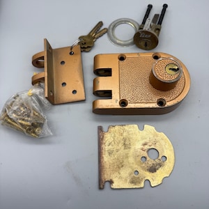 May include: A disassembled copper-colored door lock set. The set includes a main lock body, strike plate, mounting bracket, keys, and screws. The lock has a textured surface and a round keyhole.