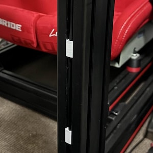 May include: Close-up of a black metal frame with white cable clips. The frame is attached to a red racing seat.