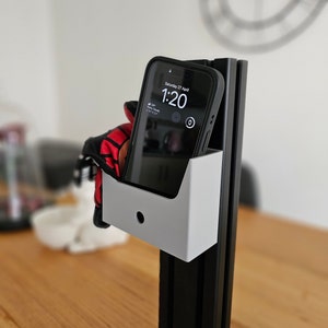 May include: A white phone holder with a black phone inside. The phone screen shows the time as 1:20 and the date as Saturday 27 April. The phone holder is attached to a black metal stand.