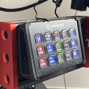 May include: A black Stream Deck with a red and black mounting bracket. The Stream Deck has a variety of buttons with labels such as "IGN", "START", "DELTA", "JASH", "ORRY", "THX", and "ARB".