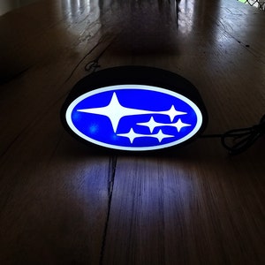 May include: A black oval sign displaying the Subaru logo, a cluster of white stars on a blue background. The sign is lit, and the logo is clearly visible. The sign is connected to a cable.