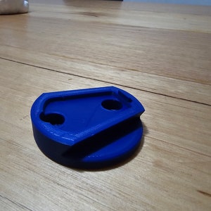 May include: A bright blue, 3D-printed item with a distinctive form. It has a circular base and a top section with two circular holes and a curved edge. The object is made of solid plastic and rests on a wooden surface.
