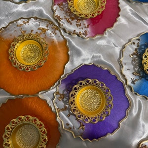 May include: A selection of colourful resin coasters with gold-coloured decorative holders. The coasters feature a variety of colours, including blue, orange, purple, and green, with gold leaf accents. Each coaster has a unique, organic shape.