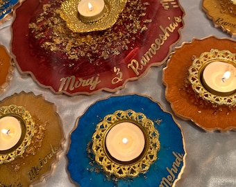 Puede incluir: Conjunto de seis posavasos de resina coloridos con detalles dorados y un pequeño portavelas en el centro de cada uno. Los posavasos están decorados con pan de oro y tienen nombres escritos en ellos, incluyendo "Miraj & Rameesha's Mehndi".