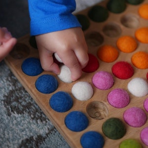 May include: A wooden board with a grid of circular indentations, each filled with a colourful felt ball. The balls are arranged in a rainbow pattern, with white balls in the centre. A child's hand is reaching into the board to pick up a white ball.