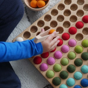 May include: A wooden counting board with numbered spaces filled with colourful felt balls. A child's hand uses a wooden tong to pick up a ball. The board is a learning tool for counting and colour recognition.