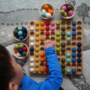 May include: A wooden counting board with 100 numbered compartments, filled with colourful felt balls. A child's hand is reaching into the board to place a ball. There are four bowls of felt balls in different colours on the table.