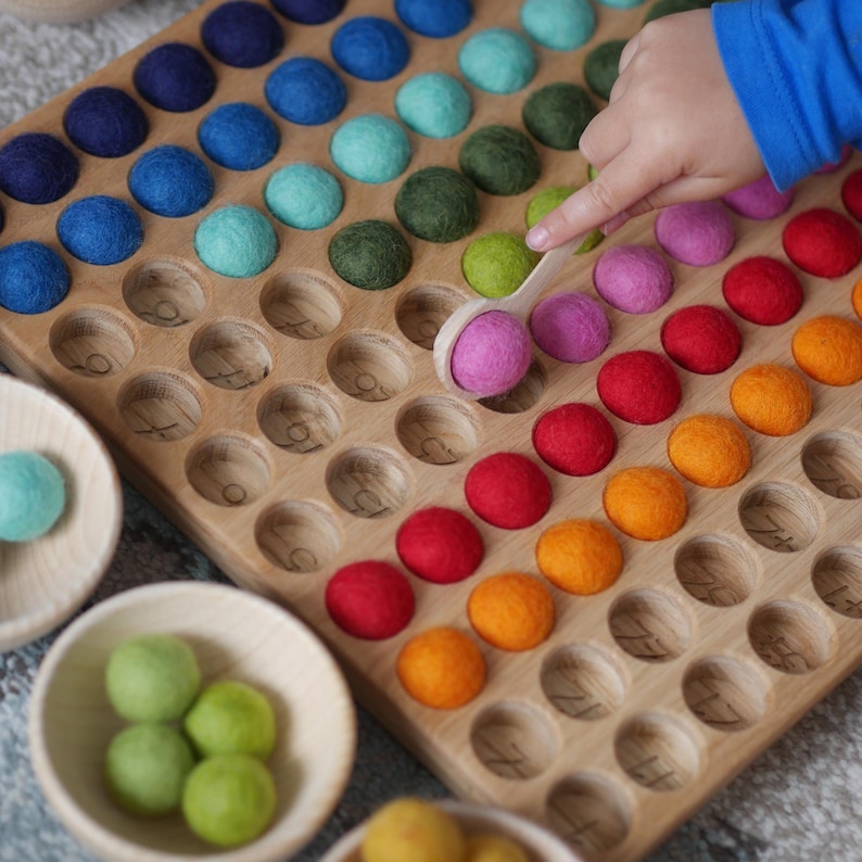 May include: A wooden board with a rainbow of felt balls in a grid pattern. A child's hand is using a wooden spoon to pick up a pink ball. The board has numbers printed on it.