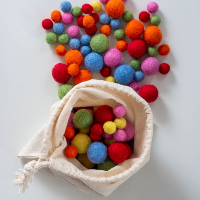 Op de afbeelding: Een witte trekkoordtas gevuld met kleurrijke viltballen. De tas ligt op een wit oppervlak. De viltballen zijn in verschillende kleuren, waaronder rood, oranje, geel, groen, blauw en paars.