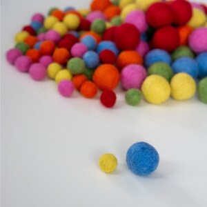 Op de afbeelding: Een verzameling kleurrijke viltballen in verschillende tinten rood, oranje, geel, groen, blauw en roze. De ballen zijn in een willekeurig patroon gerangschikt en zijn allemaal ongeveer even groot.