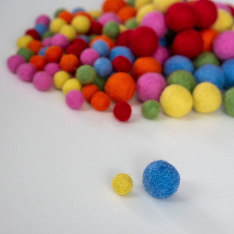 Op de afbeelding: Een verzameling kleurrijke viltballen in verschillende tinten rood, oranje, geel, groen, blauw en roze. De ballen zijn in een willekeurig patroon gerangschikt en zijn allemaal ongeveer even groot.