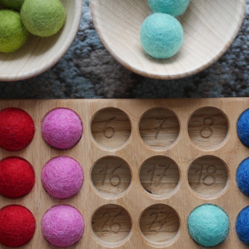 May include: A wooden tray with 18 circular compartments, each with a number inscribed. The compartments are filled with colourful felt balls in red, pink, green, and blue.