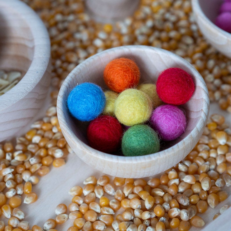 Op de afbeelding: Een houten kom gevuld met kleurrijke viltballen. De ballen zijn rood, oranje, geel, groen, blauw en paars. De kom staat op een oppervlak bedekt met ma&iuml;skorrels.