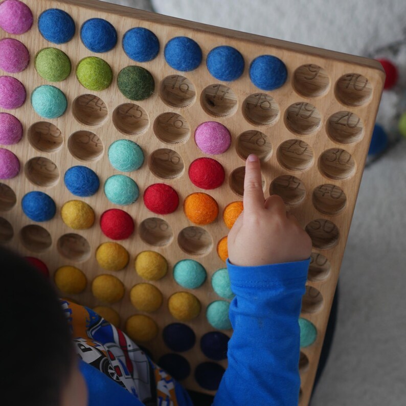 May include: A wooden board with numbered circles, each containing a colourful felt ball. A child's hand points to a circle with a pink ball. The board is a learning tool for counting and colour recognition.