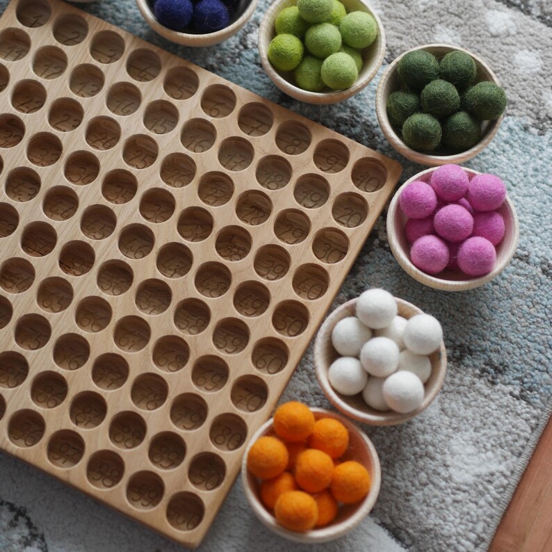 May include: A wooden counting board with numbered circles, surrounded by bowls of colourful felt balls. The bowls contain green, white, pink, orange, and blue felt balls.