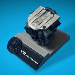 Puede incluir: Modelo detallado de un motor V8 Kompressor, principalmente plateado y negro, montado sobre una base de dos niveles. La base presenta el texto "V8 KOMPRESSOR" y un emblema circular. El modelo del motor tiene detalles intrincados.