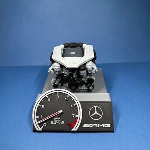 Könnte beinhalten: Ein weiß-schwarzes Modell eines Automotors mit einem schwarzen Sockel. Der Sockel hat ein silbernes Mercedes-Benz-Logo und den Text "AMG". Ein schwarz-weißes Tachometer mit einem roten Zeiger ist am Sockel befestigt. Das Tachometer zeigt "6.3-V8" und "KJ000 1/min" an.