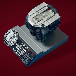 Puede incluir: Modelo plateado de un motor V8, montado sobre una base gris. El motor tiene componentes negros y el texto "AMG" es visible. También hay una placa con texto que incluye "5.4L V8 M113K" y "E 55 AMG". El fondo es rojo oscuro.
