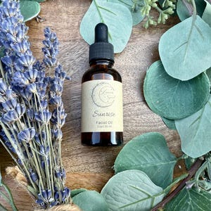 May include: A brown glass bottle with a dropper top, labeled "Sunrise Facial Oil 1 fl oz / 30 ml". The bottle is surrounded by dried lavender and eucalyptus leaves.