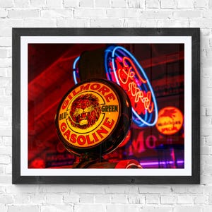 Op de afbeelding: Ingelijste print met vintage neonreclames. De focus ligt op een Gilmore Gasoline-bord met een leeuwenembleem, tegen een achtergrond van rode en blauwe neonlichten, inclusief een Ford-bord. De afbeelding heeft een retro esthetiek.