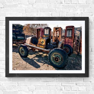 Op de afbeelding: Een ingelijste print met een vintage raceauto met nummer 8, geparkeerd voor oude benzinepompen. De auto en pompen zijn roestig en verouderd, wat suggereert dat het overblijfselen uit het verleden zijn. De scène speelt zich buiten af.