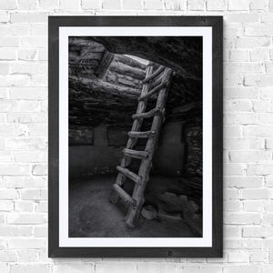 May include: Black and white framed photograph of a rustic wooden ladder leaning against a stone wall inside a dimly lit structure. The ladder leads up to an opening in the ceiling. The image is framed in a black wooden frame.