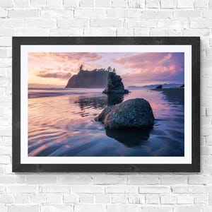 May include: A framed photograph of a coastal landscape at sunset. The image features a rocky beach with large boulders and a small island in the distance. The sky is filled with pink and purple clouds, reflecting in the wet sand.