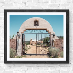 May include: A framed photograph of a church entrance with an open wrought iron gate. The archway is beige with a statue, and the sky is blue. The gate leads to a gravel path and another building in the distance.