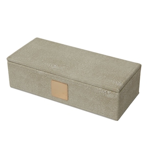 Puede incluir: Una caja rectangular de color gris claro con un pequeño cuadrado dorado en la parte superior. La caja está hecha de un material texturizado.
