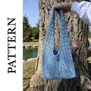 Pode incluir: Uma bolsa tote de crochê azul claro com uma alça única. A bolsa está pendurada em um tronco de árvore.