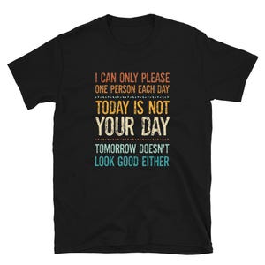 Könnte beinhalten: Schwarzes T-Shirt mit dem Text "I CAN ONLY PLEASE ONE PERSON EACH DAY, TODAY IS NOT YOUR DAY, TOMORROW DOESN'T LOOK GOOD EITHER" in orange, weiß und blau.