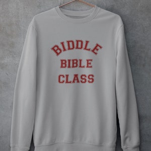 May include: A light gray sweatshirt with the words "BIDDLE BIBLE CLASS" in red, collegiate-style lettering. The sweatshirt is hanging on a copper-colored hanger against a gray concrete wall. The sweatshirt has a crew neck and long sleeves.