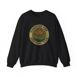Puede incluir: Sudadera negra de cuello redondo con un gráfico circular. El gráfico presenta un contenedor de basura en llamas con las palabras "DUMPSTER FIRE RESPONSE TEAM" en un diseño circular. El gráfico es en tonos marrones, verdes y naranjas.