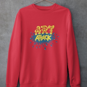 Puede incluir: Sudadera roja con un estampado gráfico amarillo que dice "ART ATTACK" con salpicaduras de pintura azul.