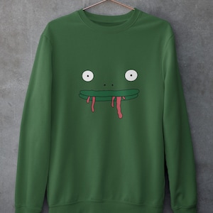 以下が含まれることがあります： 緑色のスウェットシャツに、漫画の蛙の顔のグラフィックがプリントされています。蛙は大きな白い目、緑色の口、赤い垂れ下がった舌をしています。