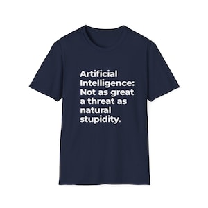 以下が含まれることがあります： 紺色のTシャツ。白い文字で「Artificial Intelligence: Not as great a threat as natural stupidity.」と書かれています。テキストは中央に配置されています。Tシャツはクルーネックで半袖です。