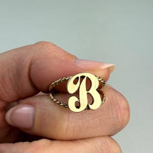 May include: A gold-colored ring featuring the letter "B" in a bold, cursive font. The ring band has a twisted rope design. The ring is shown on a person's finger against a soft, neutral background. Jewelry.