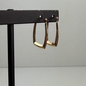 May include: A pair of gold-colored, rectangular hoop earrings. The earrings are displayed on a black earring stand against a light gray background. The earrings have a polished finish and a simple, modern design.