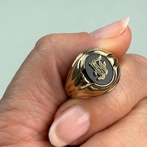 May include: A gold signet ring with a black oval center. The center features a gold monogram. The ring is held by a hand with manicured nails. The ring has a polished, reflective surface.