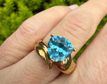 May include: A gold ring featuring a large, triangular, light blue gemstone. The ring has a unique, flowing design with a polished finish. The gemstone is securely set with small prongs, showcasing its vibrant color and clarity. A piece of jewelry.